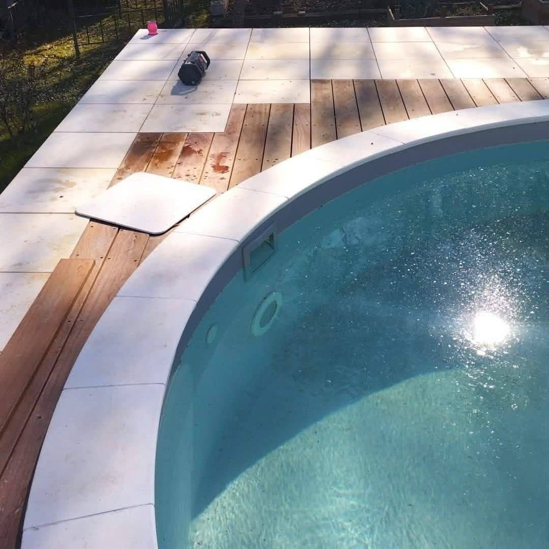 Vue plongeante sur une piscine à plage mixte, dalles claires et lames de bois exotique, entourée d’un jardin arboré.