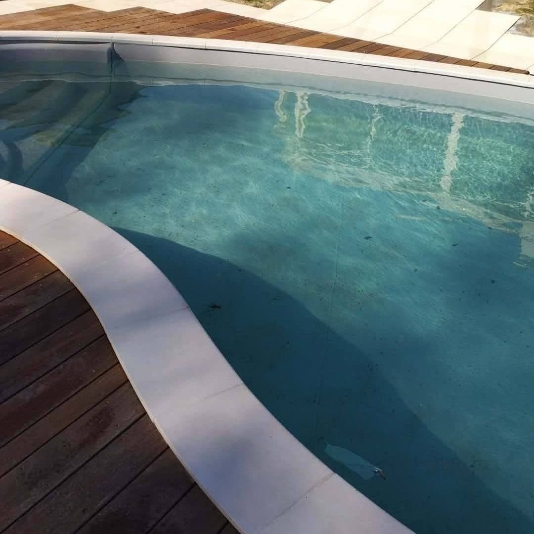 Piscine et terrasse en bois et dallage clair aménagées au pied d’une maison, avec baie vitrée et mobilier de jardin en arrière-plan.